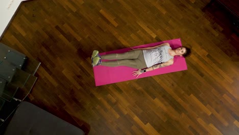 Top-down-view-of-a-tattooed-woman-doing-leg-raises-on-a-pink-yoga-mat,-indoor-fitness-concept