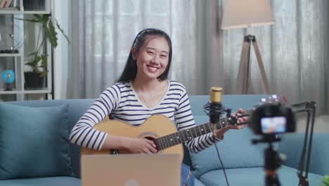 mujer asiática con una computadora portátil sosteniendo una guitarra y sonriendo a la cámara mientras transmite en vivo en casa