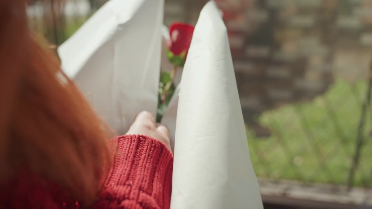 Primo piano di una donna con i capelli rossi che indossa un maglione di lana, mentre scarta con cura della carta bianca per rivelare dei fiori. La scena è ambientata all'aperto, alla luce naturale del giorno, con un focus sull'interazione tattile.