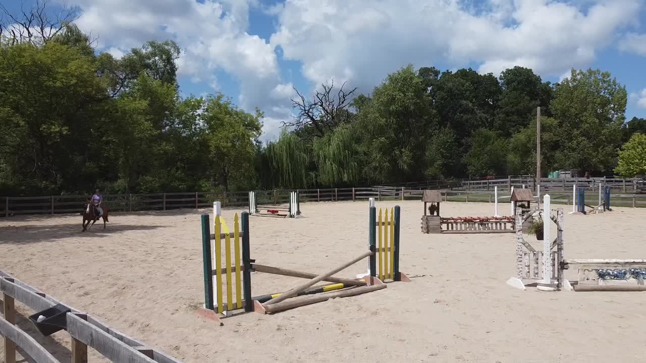 Uma menina em um pônei cavalga por uma arena de equitação ao ar livre e salta múltiplos obstáculos.
