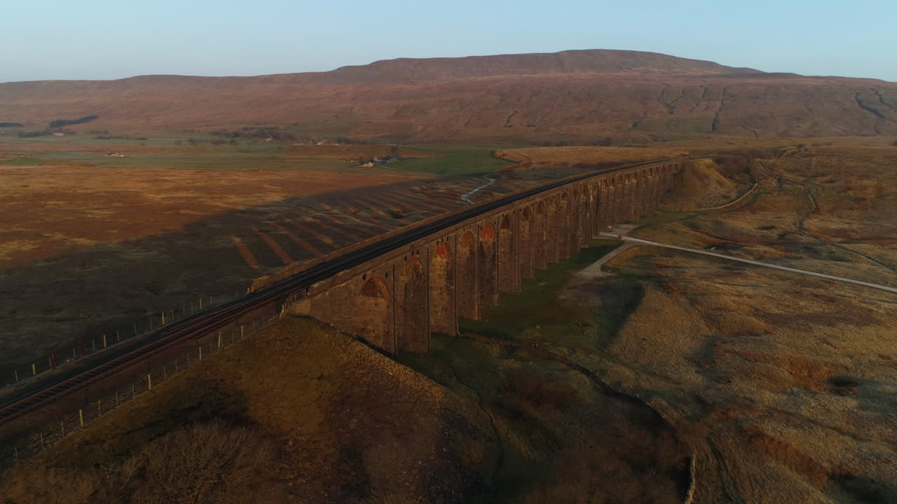 toma aérea lenta de un dron del puente del tren del viaducto ribblehead con largas sombras en el impresionante amanecer en verano en los valles de yorkshire inglaterra reino unido con 3 picos de la montaña whernside en el fondo