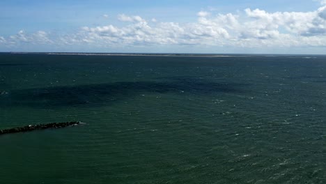 Ein-Zeitraffer-Vom-Strand-Von-Coney-Island,-An-Einem-Sonnigen-Tag,-Während-Der-Blick-Der-Drohnenkamera-Auf-Den-Atlantik,-Den-Horizont-Mit-Blauem-Himmel-Und-Ein-Paar-Weißen-Wolken-Gerichtet-Ist