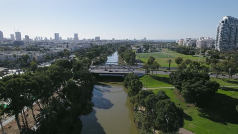 Volando-Sobre-Un-Puente-Urbano-Con-Autos-Cruzando-Un-Río-Con-Kayaks,-Amplios-Jardines-Y-Un-Campo-De-Fútbol-En-Un-Barrio-Residencial