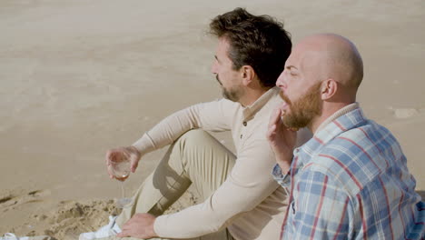 feliz pareja gay bebiendo vino y comiendo croissants mientras se sienta en la playa