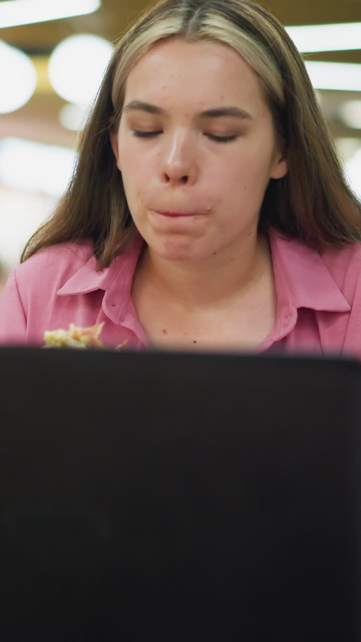 mujer de vestido rosa trabajando en una computadora portátil mientras come una hamburguesa en una mesa en un centro comercial ocupado, una taza de café está colocada al lado de la computadora portatil, y ella está enfocada en su tarea, multitarea entre el trabajo y la comida