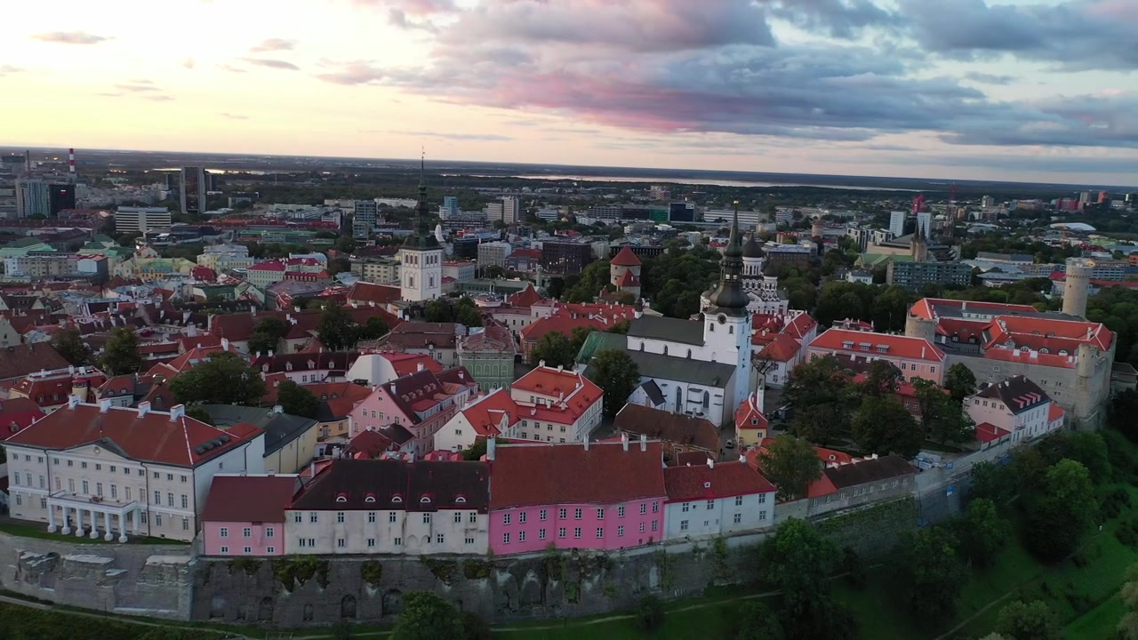 Impresionantes imágenes de dron del casco antiguo medieval de Tallin al atardecer. La toma muestra la colina de Toompea, la Catedral de Santa María y edificios con tejados rojos bajo un cielo dramático de nubes rosas y naranjas.