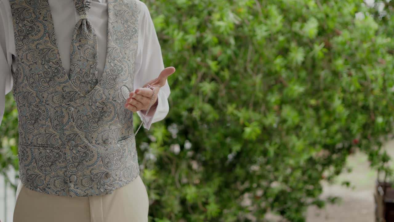 Groom standing in a detailed vest, gesturing with his hand against greenery backdrop