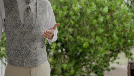 groom standing in a detailed vest, gesturing with his hand against greenery backdrop