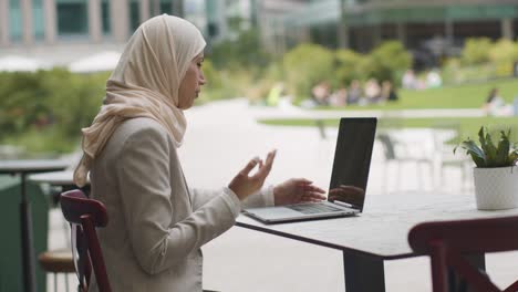 Muslim-Businesswoman-Sitting-Outdoors-In-City-Gardens-Making-Video-Call-On-Laptop-1