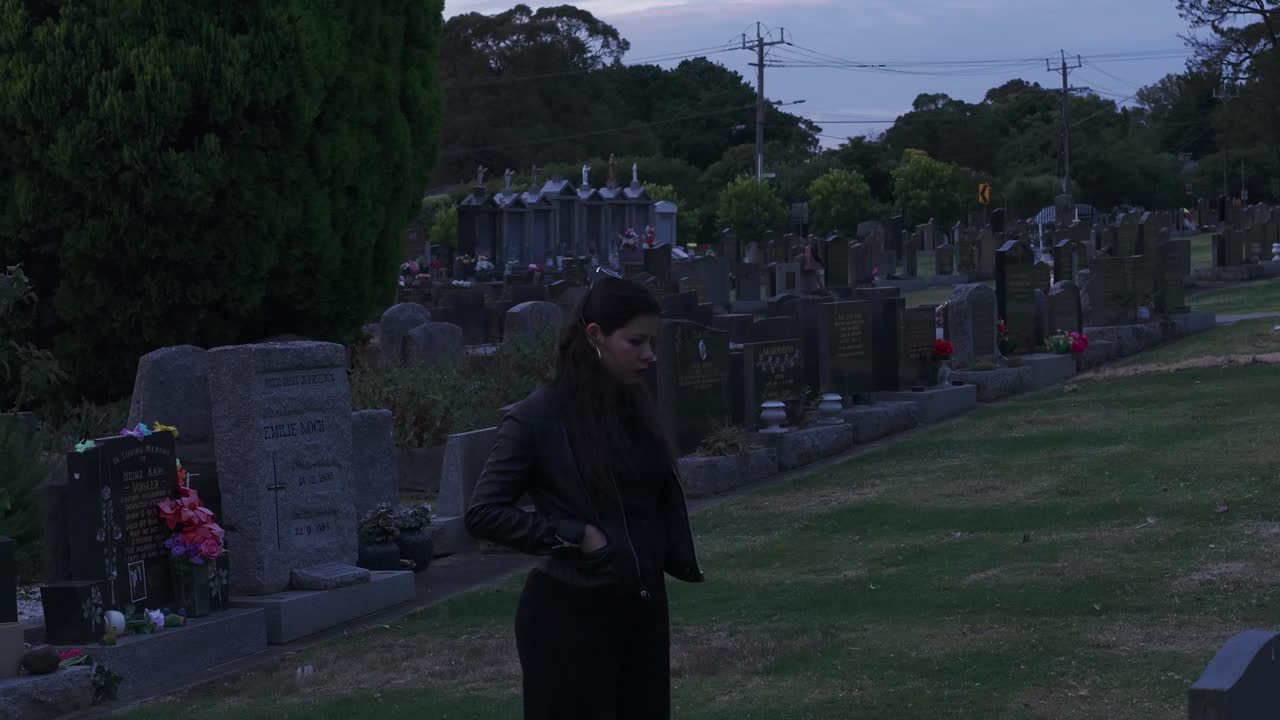 Una donna sta pensierosa in un cimitero di Melbourne mentre il sole tramonta, proiettando un tenue bagliore sulle lapidi. L'angolazione jib aggiunge profondità alla scena solenne