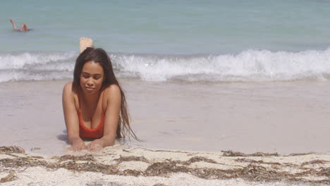 latin girl on her front in exotic beach shore shallows, plays with sand and smiles as waves gently crash, facing camera