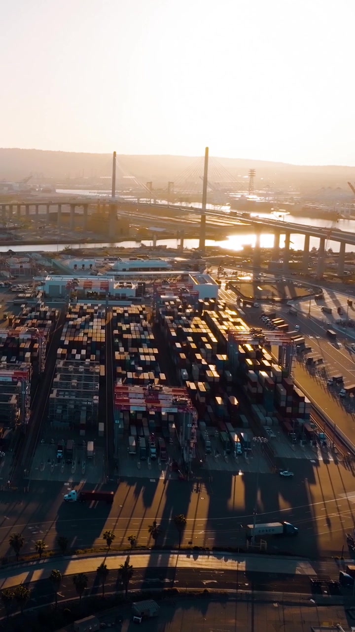 Aerial view of cargo terminal in Los Angeles port. Freight containers organized in rows. Transportation activity occurring in container yard. Sunlight reflects on waterway.