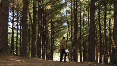 A-man-in-a-gold-jacket-walking-through-a-forest-holding-a-white-frisbee-golf-disc-in-his-hand-in-jacobson-park-lexington-Kentucky