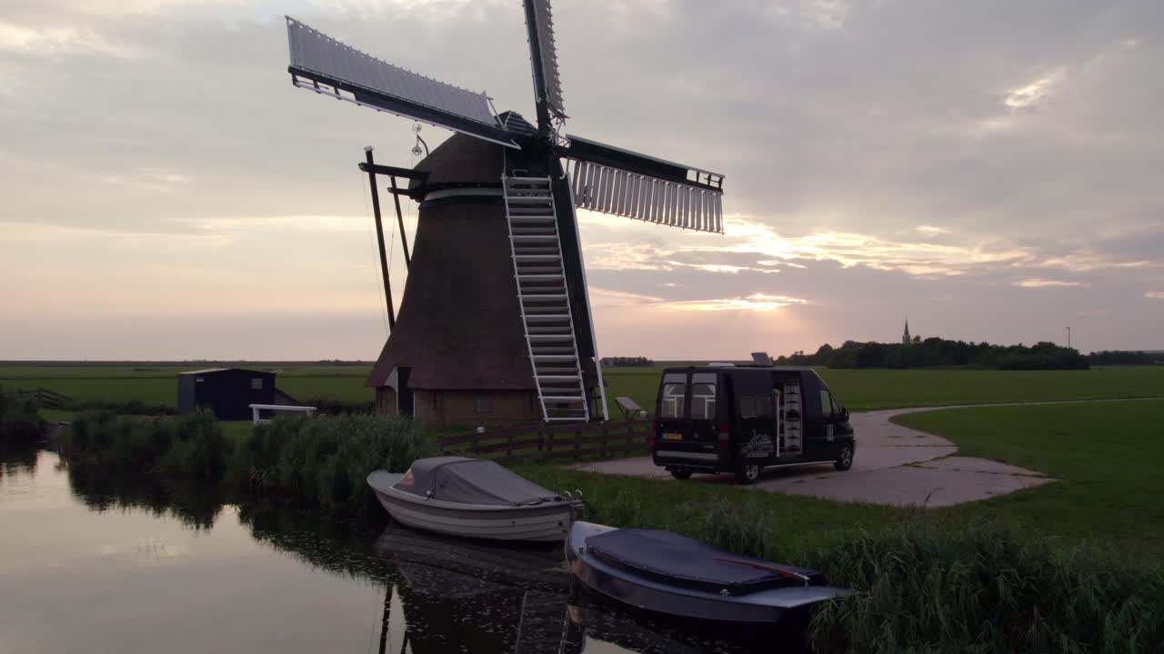 Vista aerea del mulino a vento Cornwerder Molen con camper al tramonto vicino al fiume a Cornwerd, Paesi Bassi, con illuminazione mozzafiato al tramonto, atmosfera serena, riprese drone di alta qualità che catturano momenti sereni