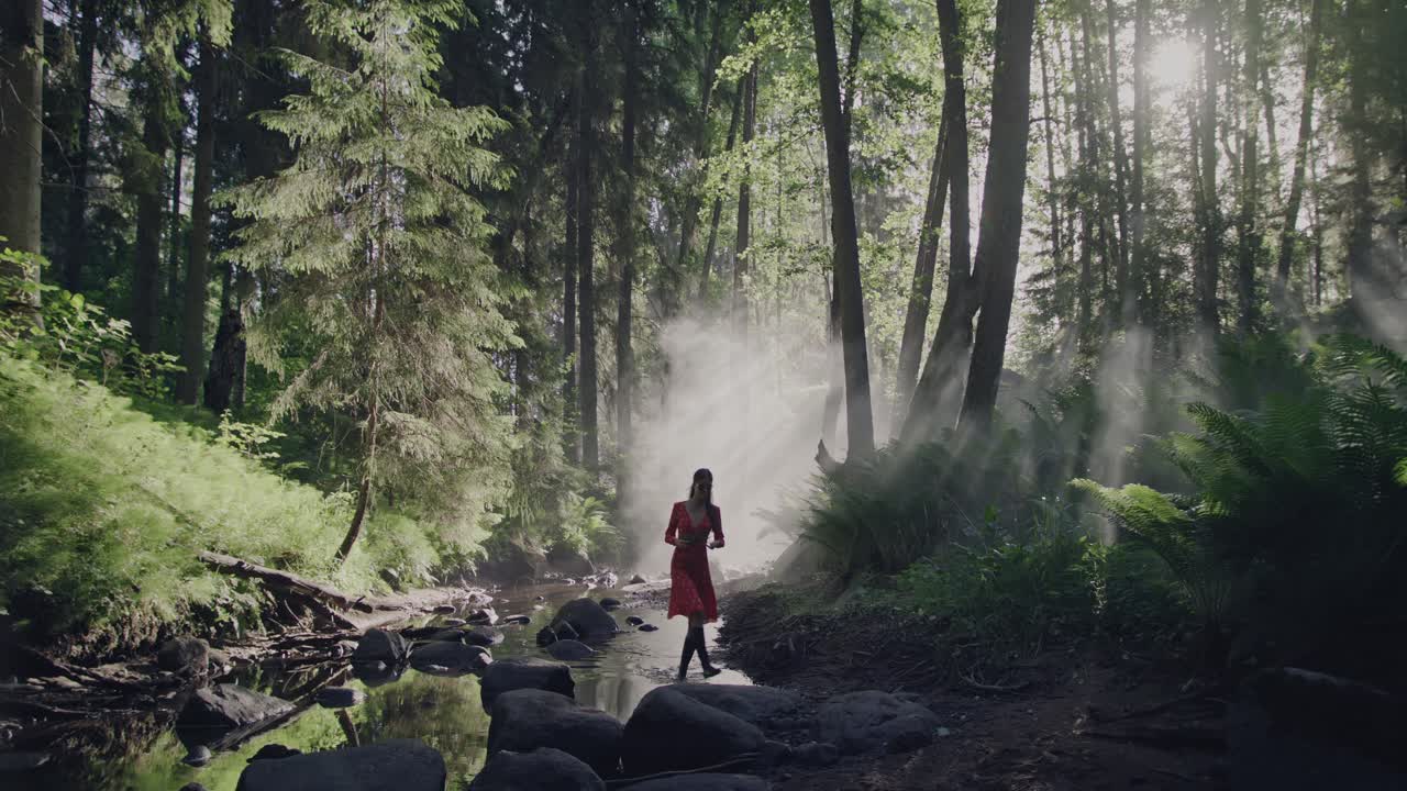 Verzauberter Waldspaziergang: Eine Frau in einem leuchtend roten Kleid geht durch einen üppigen, märchenhaften Wald entlang eines ausgetrockneten Flussbettes inmitten des mystischen Morgennebels und Dunstes.