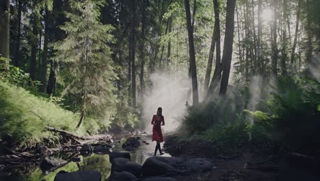 Enchanted-Forest-Stroll:-A-woman-in-a-vibrant-red-dress-walks-through-a-lush,-fairy-tale-forest-along-a-dried-riverbed-amid-the-mystical-morning-mist-and-fog