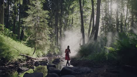 enchanted forest stroll: a woman in a vibrant red dress walks through a lush, fairy-tale forest along a dried riverbed amid the mystical morning mist and fog