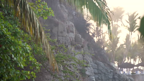 Cierre-El-Follaje-Y-El-Paisaje-De-Una-Playa-Tropical-Con-Una-Escarpada-Pared-De-Roca-Acantilado-Y-Varias-Palmeras-En-La-Costa-Arenosa