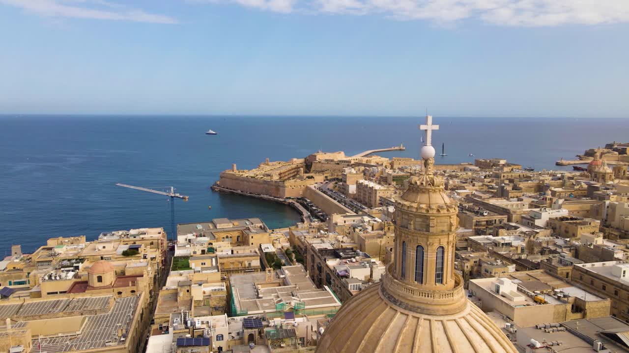 Sweeping aerial drone shot over Valletta's tightly packed golden limestone buildings, flat rooftops, and narrow streets stretching toward the Mediterranean coast and blue horizon under clear sunny ski