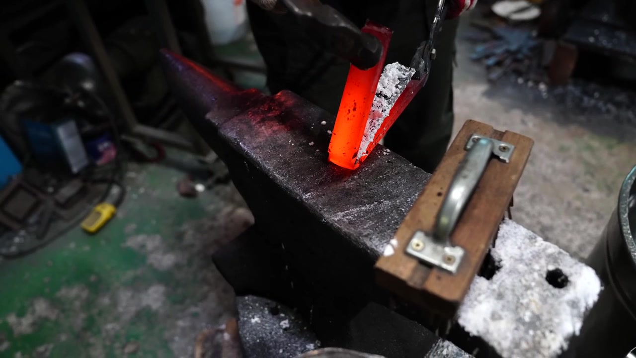 Hand hammer striking a red hot blade blank on an anvil with borax flux scattered across the work surface
