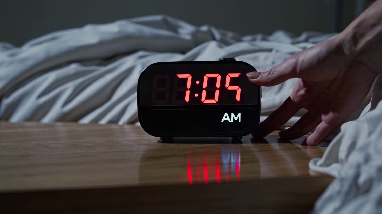 Video ravvicinato di una sveglia digitale che mostra le 7:05 del mattino su un comodino di legno, con lenzuola bianche stropicciate