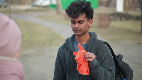 Young man with curly hair wearing gray hoodie and jeans holding orange gloves while standing in park with backpack, facing woman in pink winter jacket on early spring day