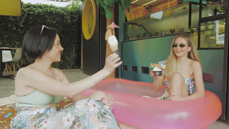 two young women enjoy ice cream in a relaxed atmosphere next to an inflatable pool in front of a contemporary ice cream parlor.
