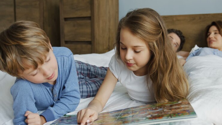 Cute little kids lying in front of the camera on the bed and reading a book for kids while their parents lying behind them in the morning