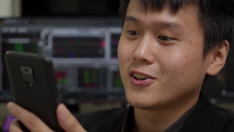 Young-Asian-man-smiling-while-looking-at-smartphone,-office-environment-with-stock-market-charts-in-background