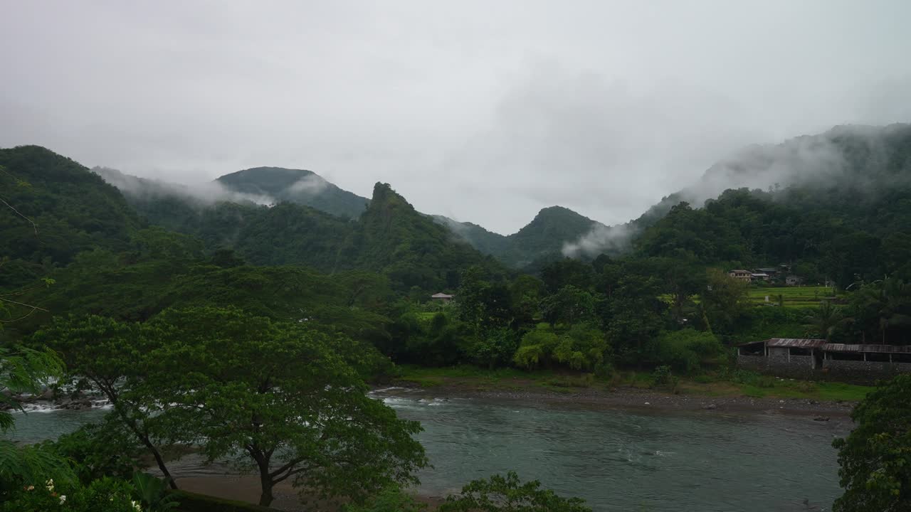 Framed from a riverside angle, lush Philippine trees line the water’s edge as mist drifts over forested mountains, with scattered homes nestled in the tranquil valley below