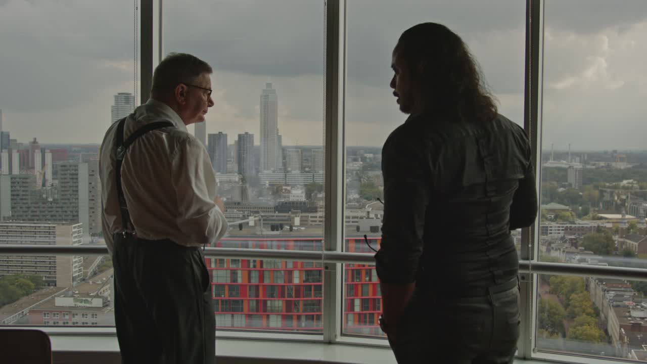 An older CEO and his employee stand in front of an office window overlooking a city skyline. The employee points to something off screen.