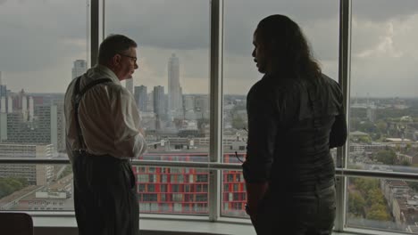 an older ceo and his employee stand in front of an office window overlooking a city skyline. the employee points to something off screen.