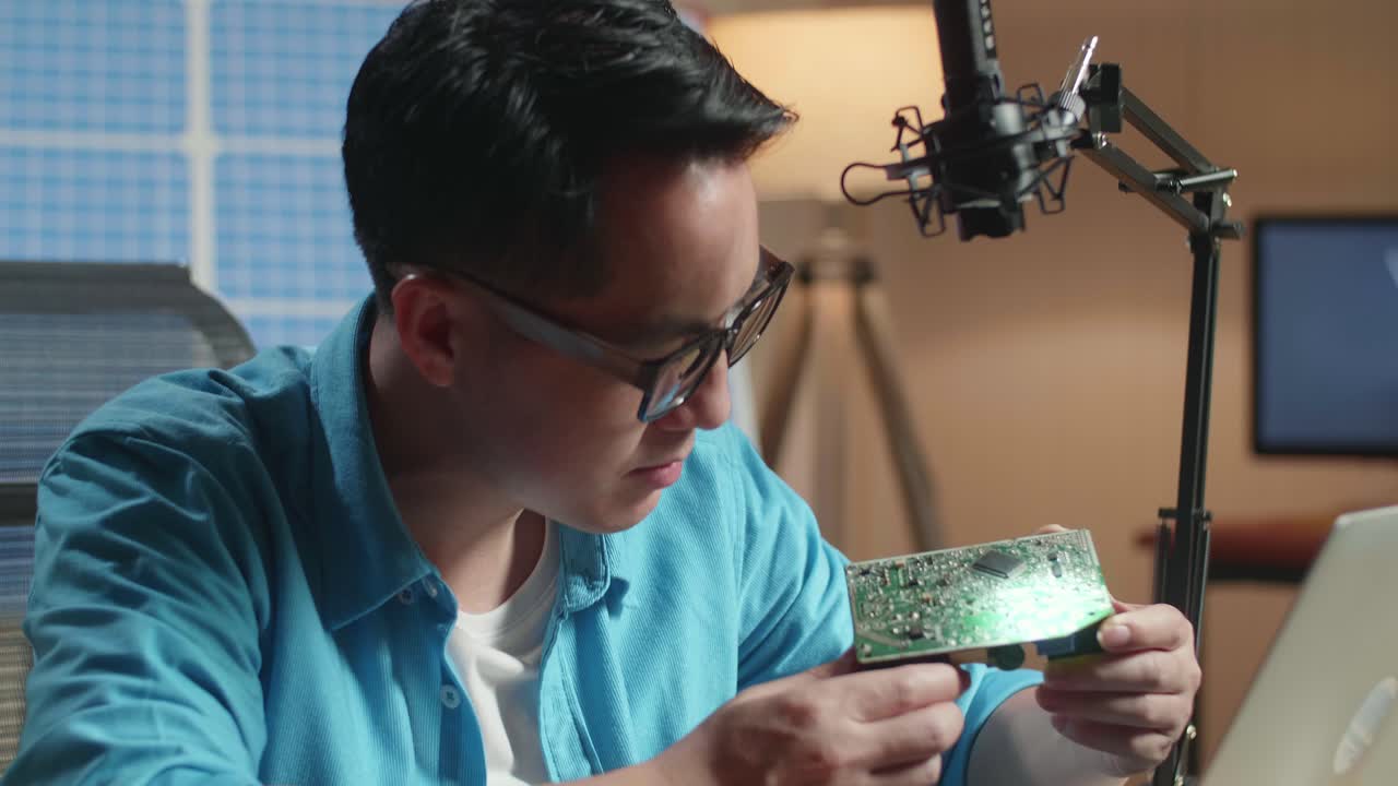 아시아인 남자가 사무실에서 노트북으로 일하면서 태양전지 앞에 앉아서 회로판을 보고 있다.