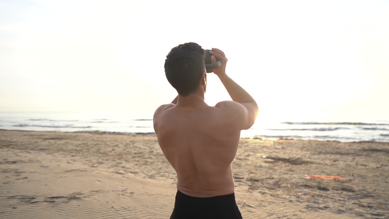 슬로우 모션으로 일출 해변에서 케틀벨과 함께 셔츠를 입지 않은 남자 훈련