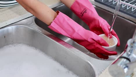 hands wearing pink gloves wash dishes in a kitchen sink under running water, focusing on cleanliness and routine