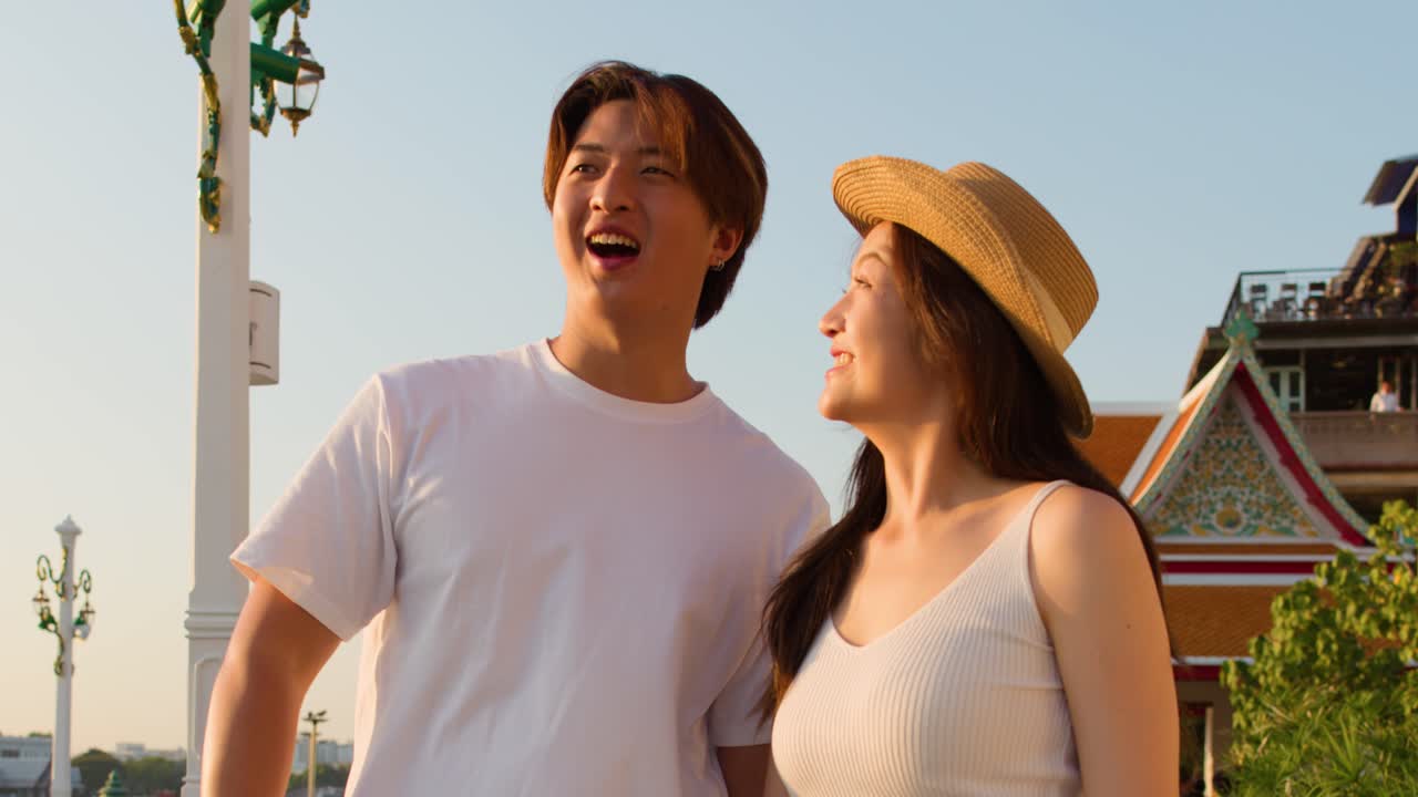 Smiling Asian couple points and admires cityscape, warm sunset lighting, temple background, handheld camera