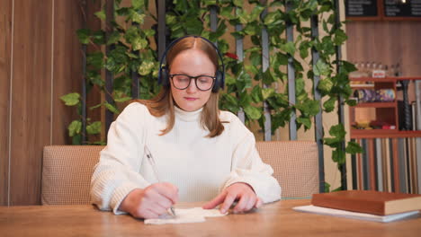 young lady wearing headphones sits at wooden table writing on napkin with pen while listening to music, background features decorative plants and blurry bar area with shelves and coffee items