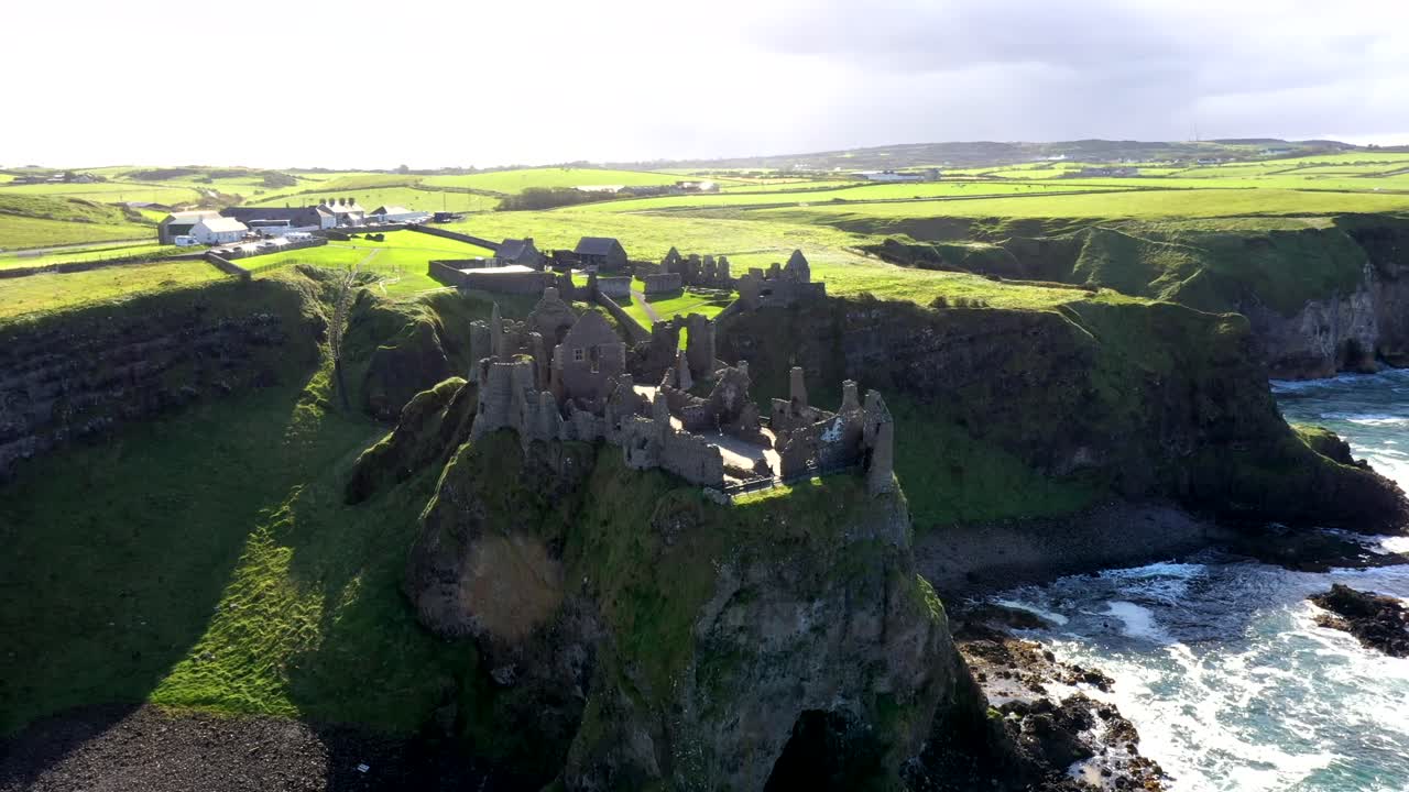 High aerial orbit of Dunluce castle on Ireland's dramatic cliffside and crashing waves, establishing overview