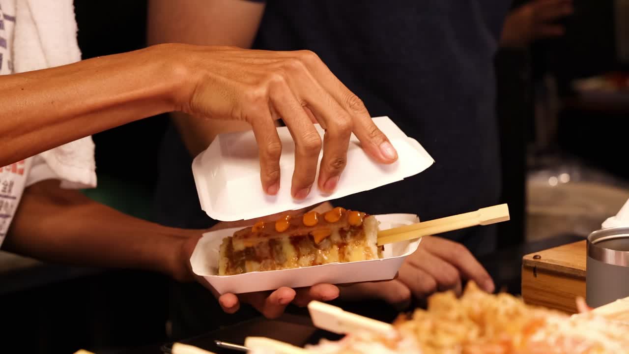 Hands prepare and serve grilled skewers with sauce, using paper trays and chopsticks.