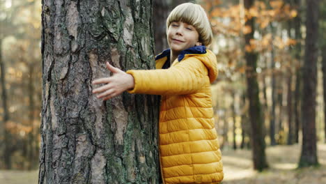 vista lateral de un lindo niño adolescente abrazando un tronco de árbol con los ojos cerrados en el bosque