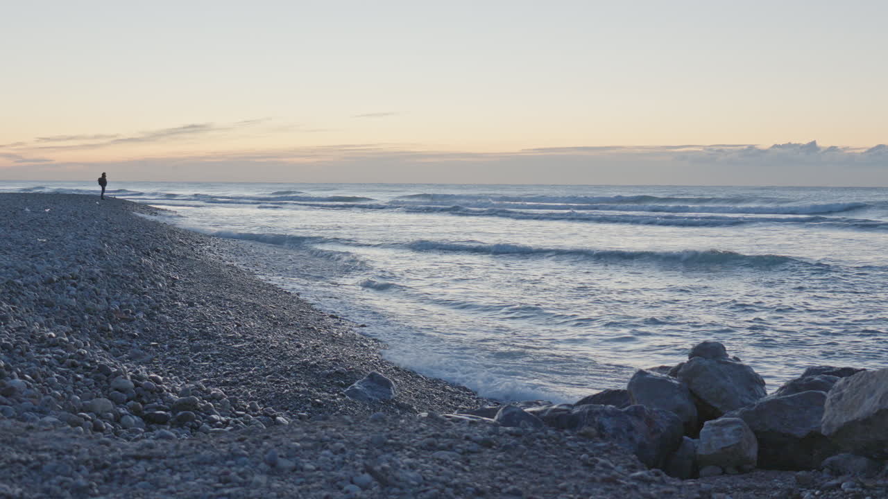 길고 돌이 많은 해변에 한 남자가 서서 파도가 들어오는 것을 지켜보고 있다