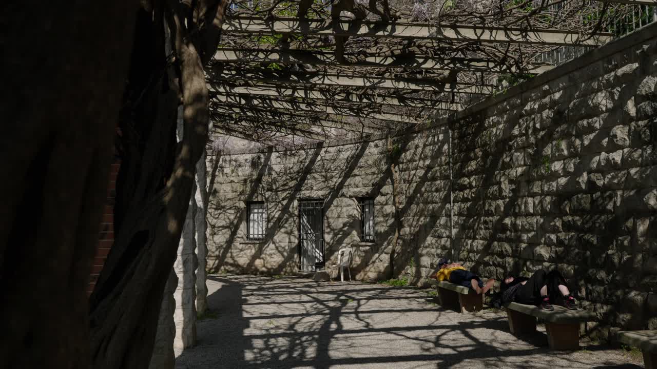 люди, отдыхающие на скамейках в историческом саду с цветами вистерии над перголами