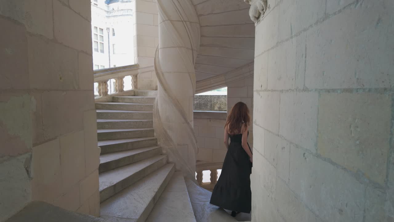 Slow motion video of a woman in a black dress descending the spiral staircase of Château de Chambord, France, inside its elegant Renaissance architecture