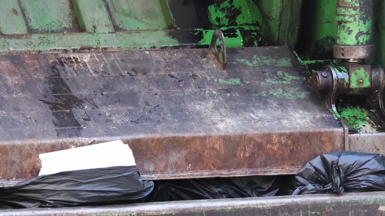 Detailed view of black trash bags being loaded into a green garbage compactor.