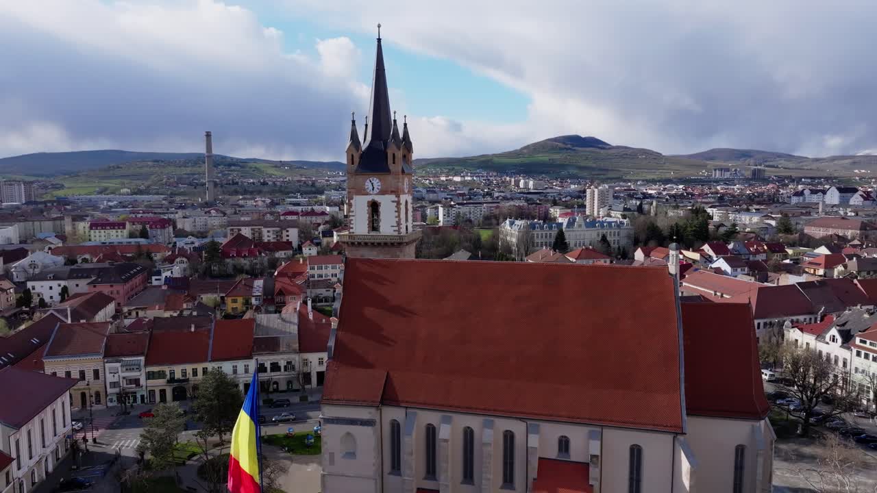 Ripresa aerea della torre rinascimentale e gotica della Chiesa Evangelica di Bistrita. La telecamera si muove fluidamente, concentrandosi sul quadrante dell'orologio, sulla guglia alta e sul paesaggio urbano circostante della Transilvania