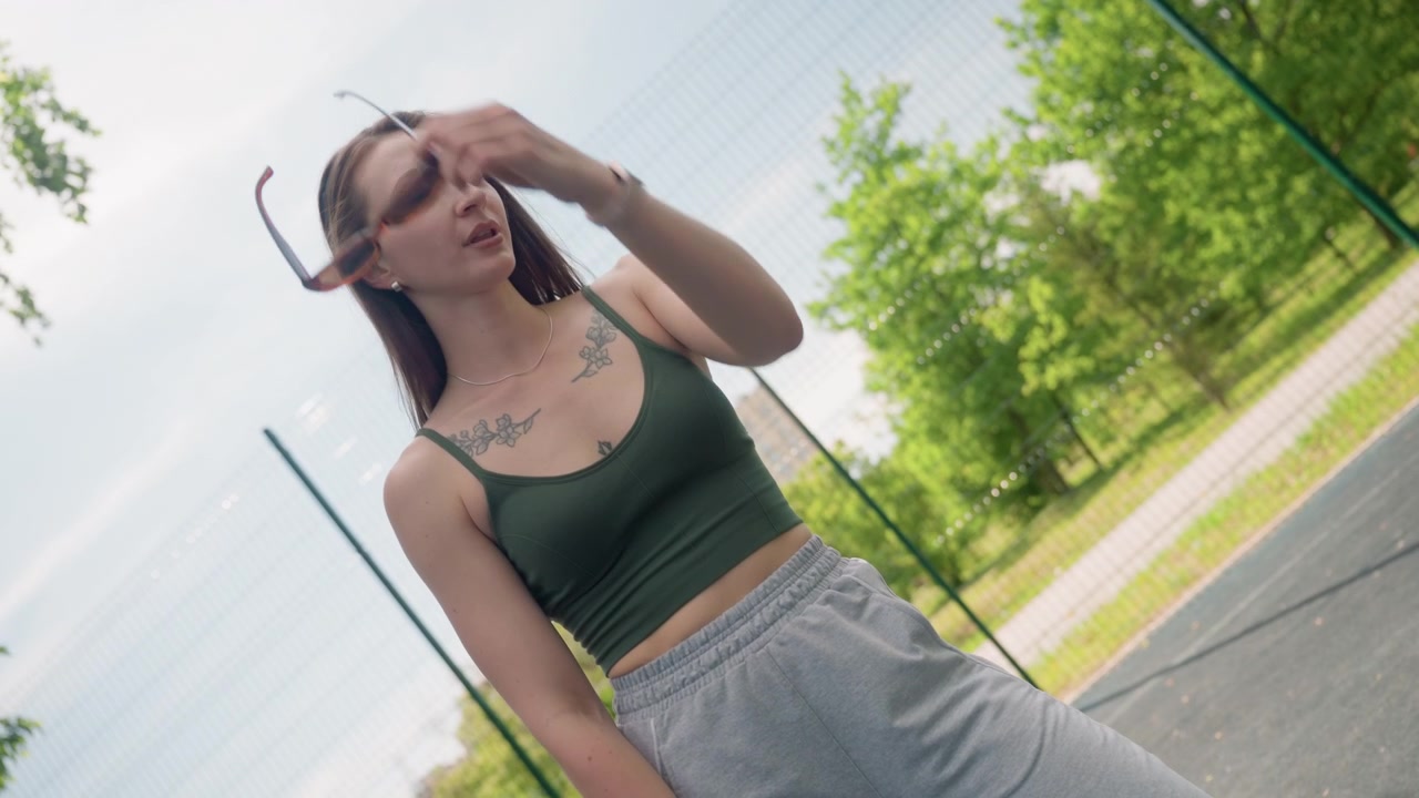 Primo piano di una giovane donna con tatuaggi che si sistema gli occhiali da sole all'aperto su una superficie asfaltata. La donna indossa un reggiseno sportivo verde e pantaloni della tuta grigi, accessoriata con orecchini, collana e smartwatch.