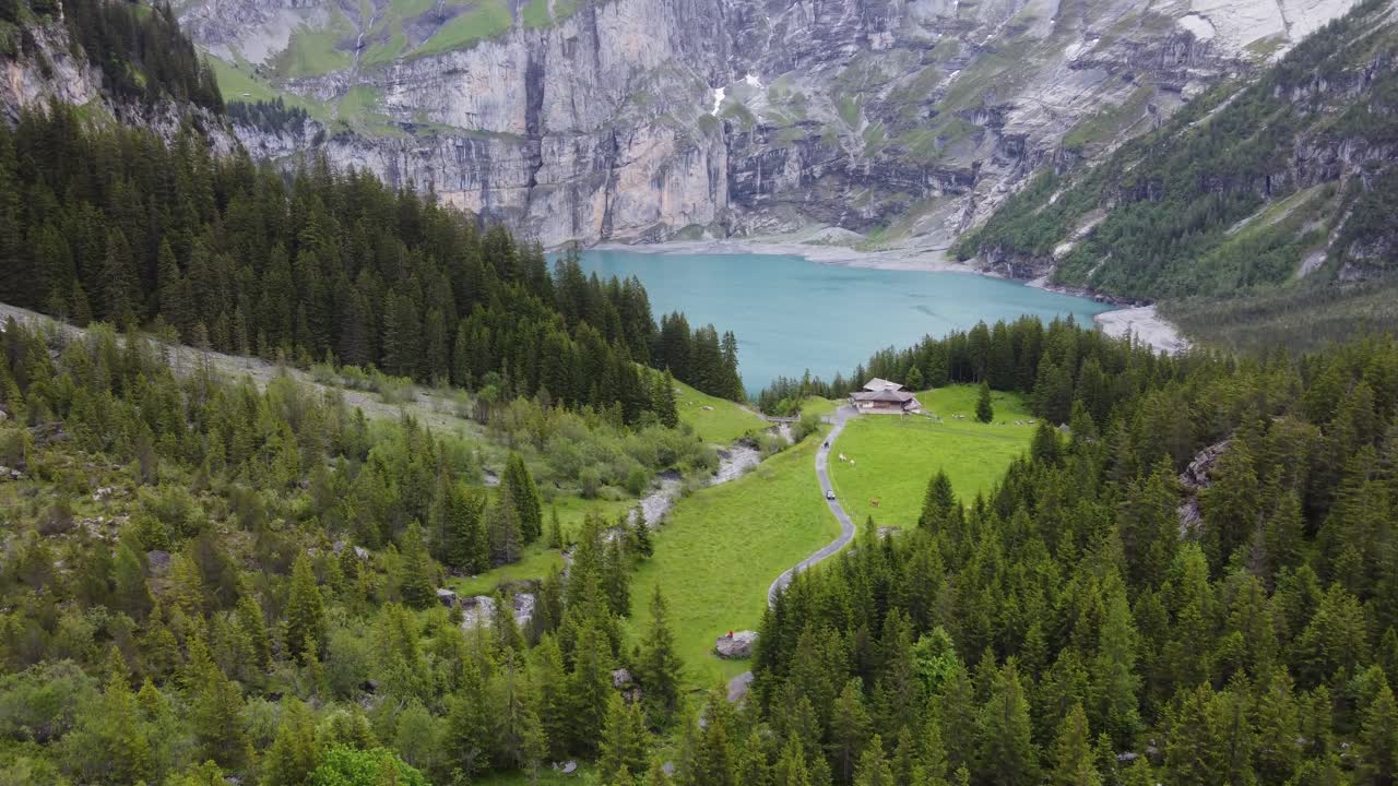 Vista aerea di una baita in legno isolata su un verde prato alpino, circondata dalle montagne e da pini che si affacciano sul lago glaciale turchese e azzurro Oeschinensee a Kandersteg, Svizzera