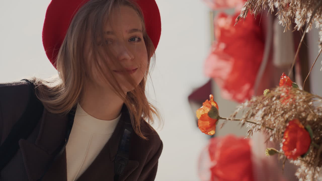 Momento romantico in una fiorista: donna che sorride e si china sui bouquet, berretto rosso e capelli mossi dal vento, assaggio giocoso di profumi e calda luce del sole, primi piani in stile influencer ed espressione gentile e gioiosa.