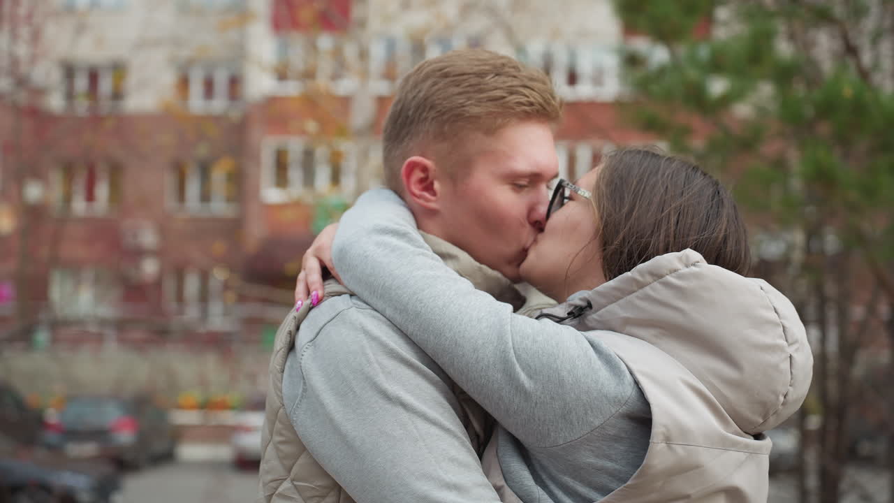 Couple bonding as they hug each other affectionately outdoors while sharing kiss in matching outfit, with woman hair blowing gently in breeze, parked cars and buildings softly blurred in background