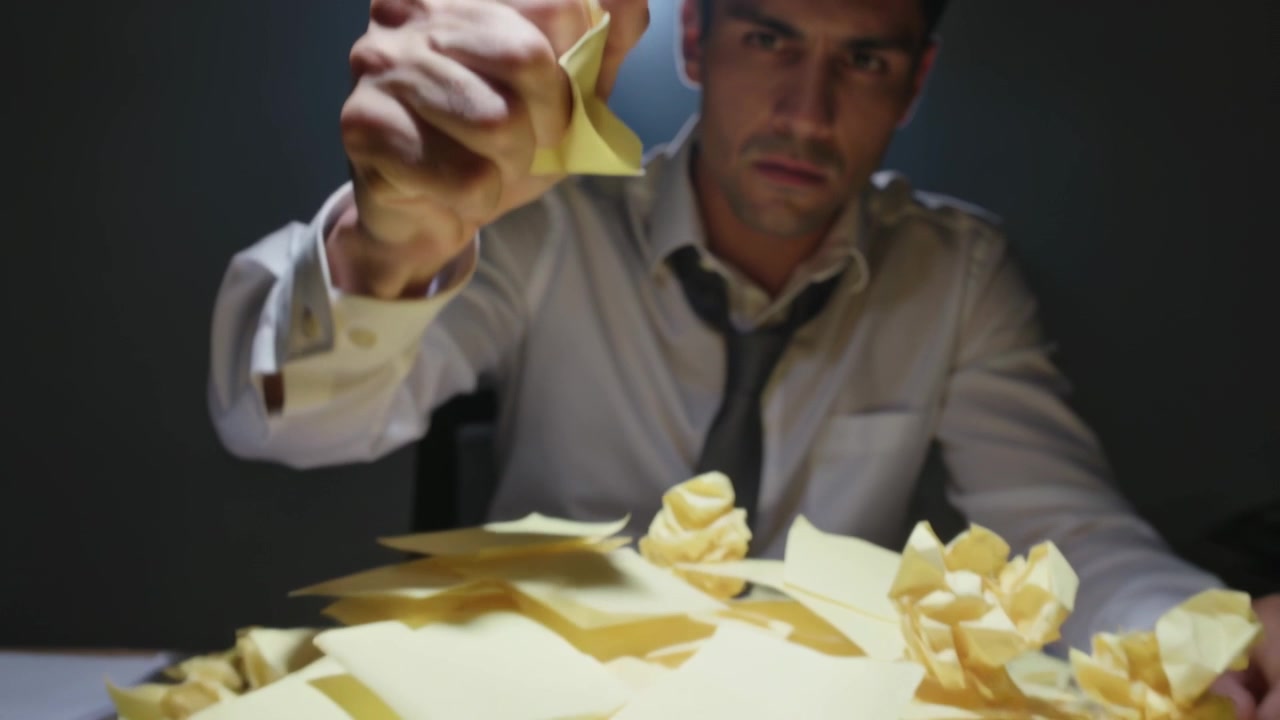 Uomo con cravatta allentata che ispeziona un biglietto giallo, piega e stropiccia appunti al tavolo sotto la lampada, controllando i documenti.